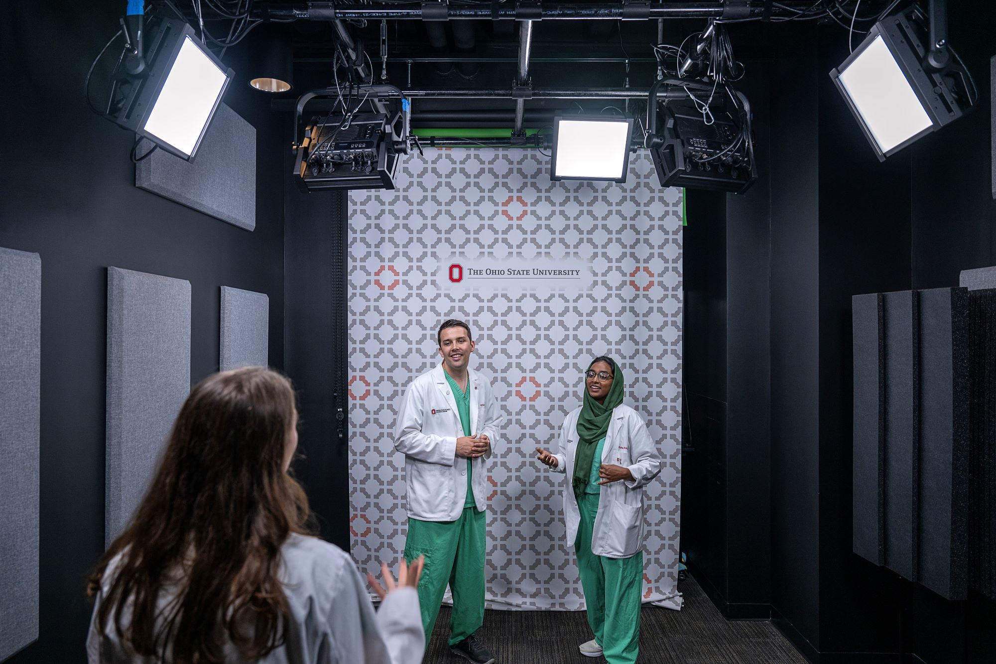 Two students in lab coats and scrubs, standing in front of an Ohio State University themed backdrop, surrounded by studio box lights and another woman in the foreground providing directions for a scene.