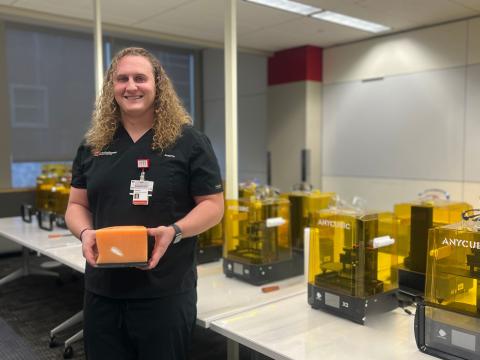 Joshua Krech standing and smiling in front of a row of 3D printers, holding a 3D printed cube.