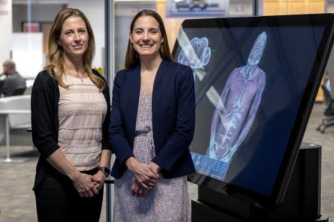 Kristen Roberts and Laura Boucher standing and smiling in front of a large digital interactive display of human anatomy.